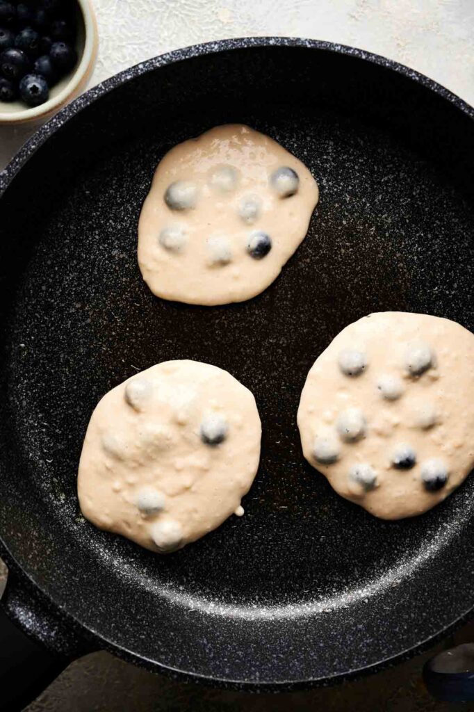 Three blueberry pancake batters cooking in a black skillet, with a bowl of blueberries partially visible in the corner.