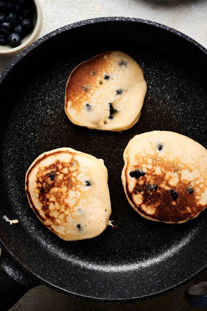 Three blueberry pancakes cooking in a black frying pan, with a bowl of blueberries partially visible in the corner.