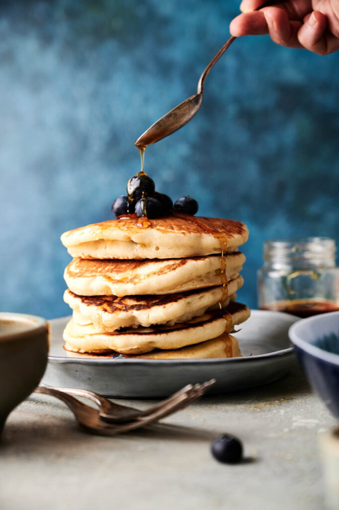 A hand drizzles syrup over a stack of pancakes topped with blueberries on a plate, with jars and utensils in the background.