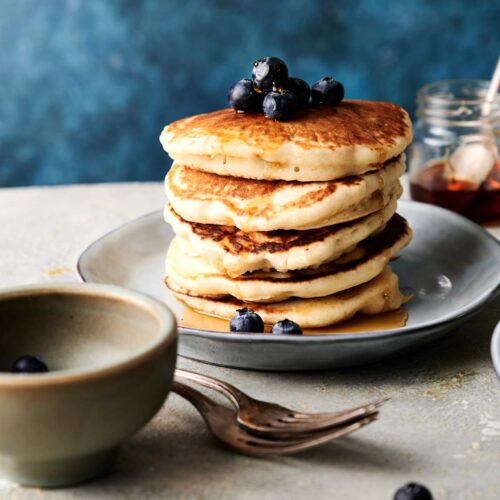A stack of pancakes topped with blueberries on a plate, with forks, a bowl, and jars of syrup nearby.