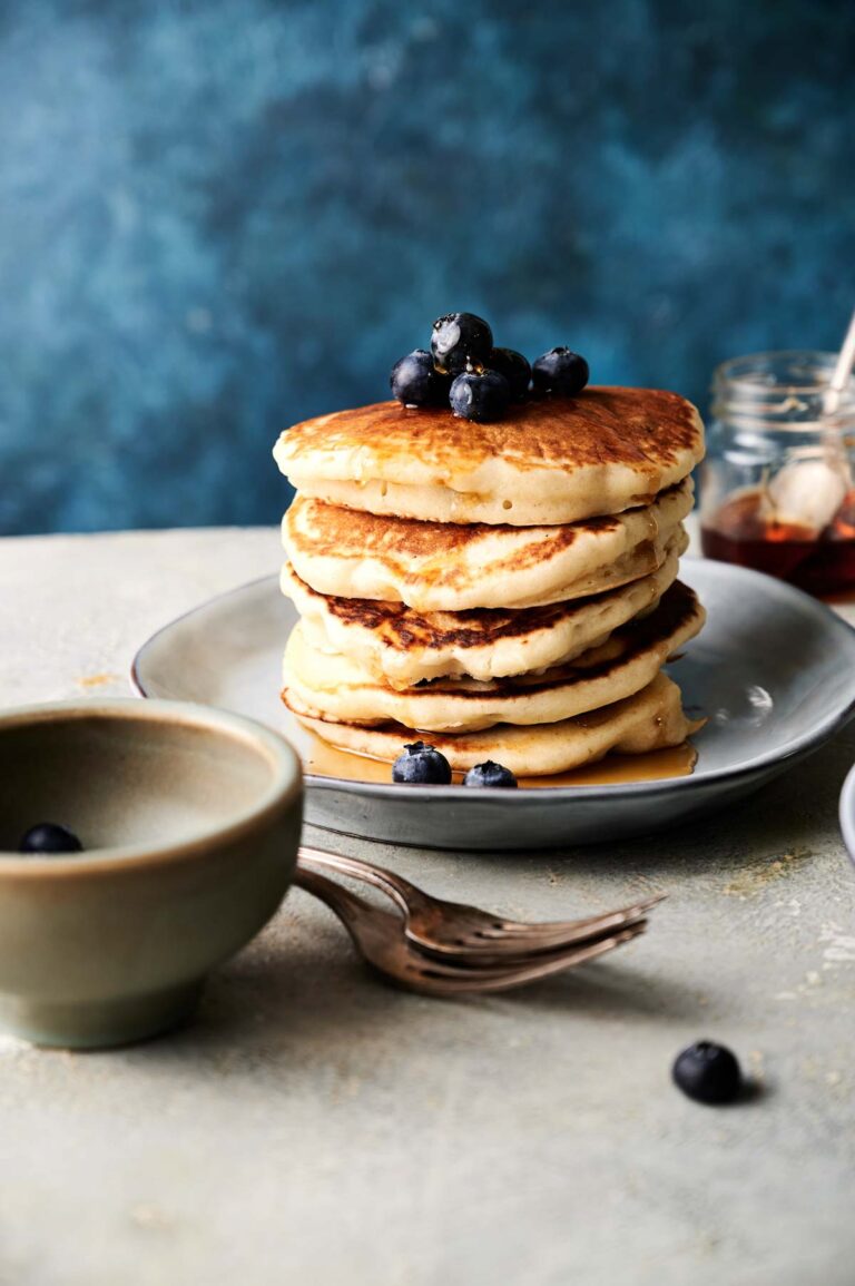 A stack of pancakes topped with blueberries on a plate, with forks, a bowl, and jars of syrup nearby.