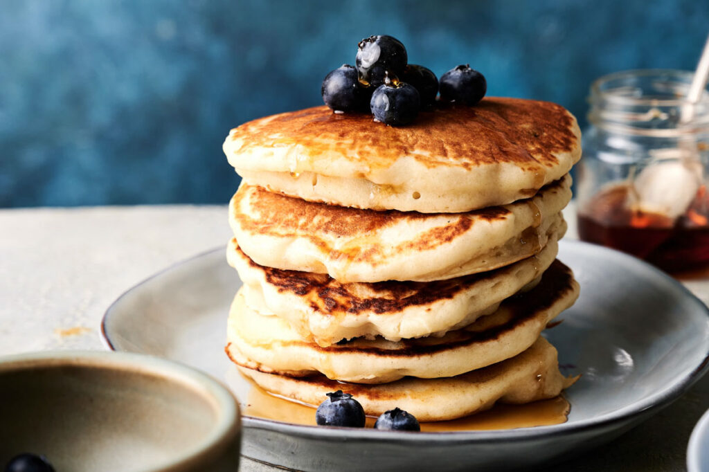 A stack of pancakes topped with blueberries and syrup on a gray plate, with a jar of honey and a bowl in the background.