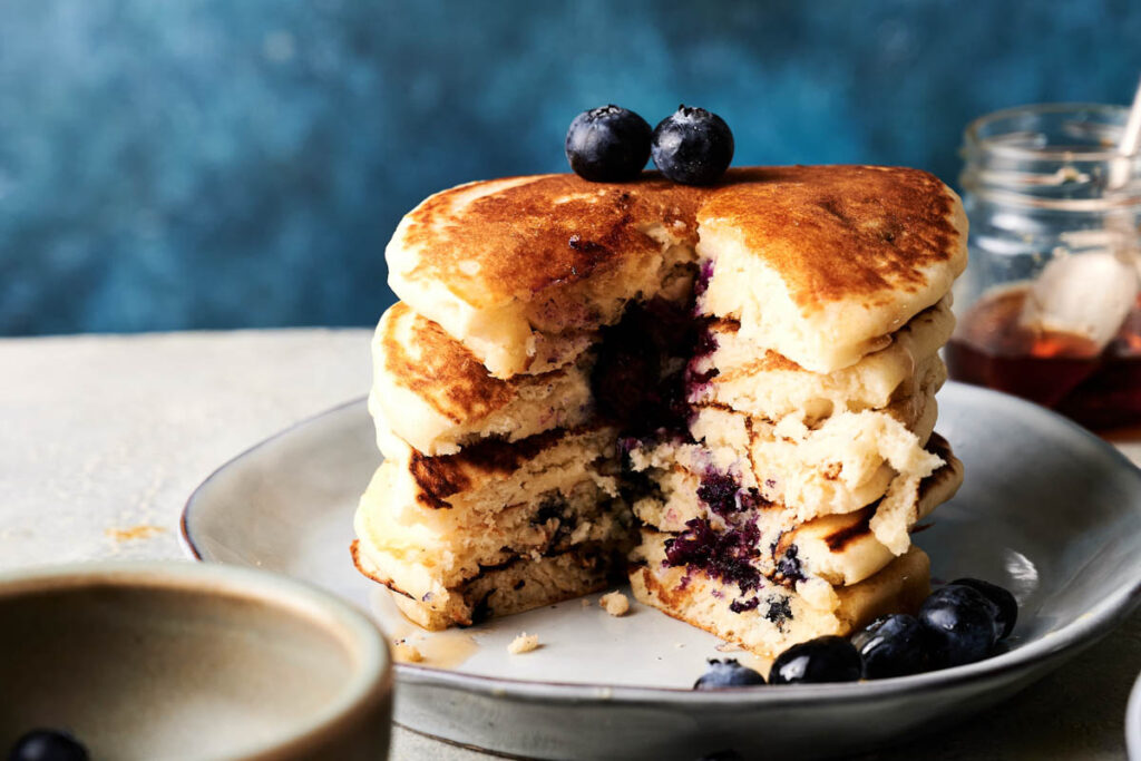 A stack of blueberry pancakes with a portion cut out, topped with whole blueberries, on a plate with a jar of syrup and a cup nearby.