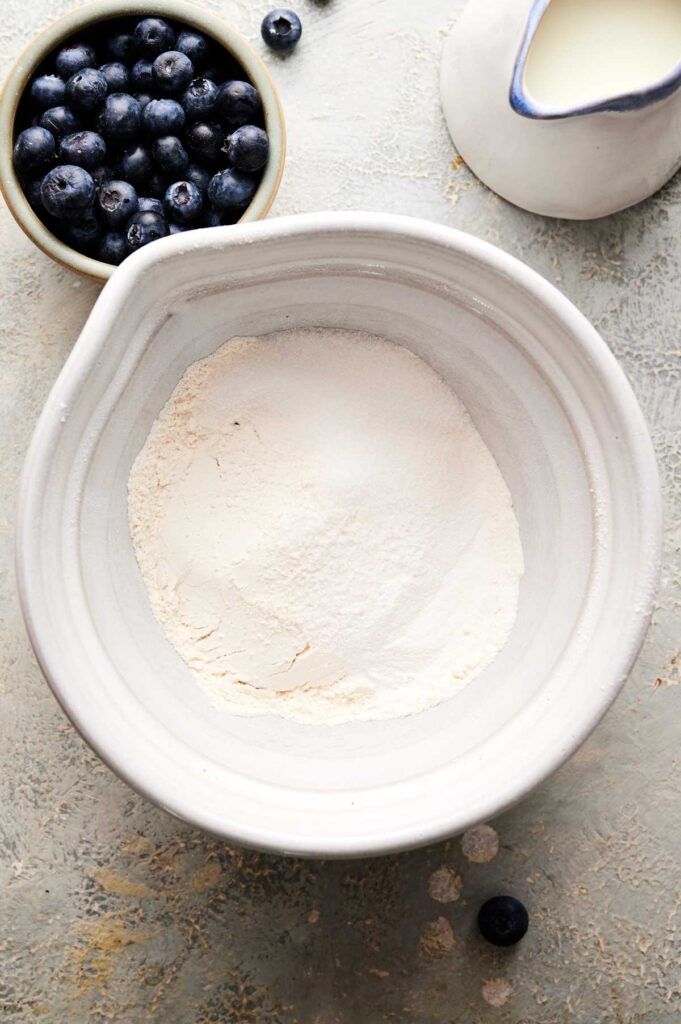 A white bowl with flour, a small bowl of blueberries, and a white pitcher of milk on a light textured surface.