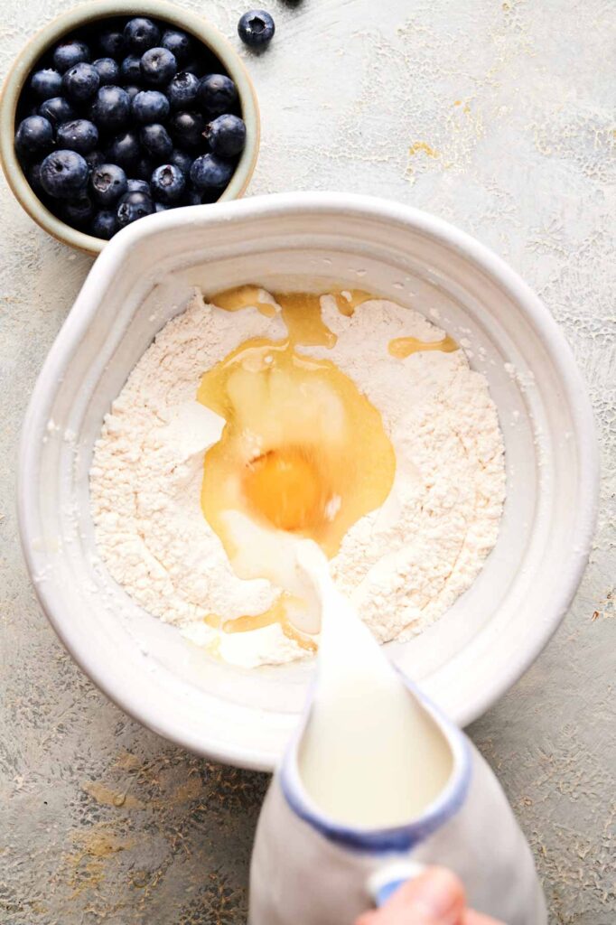 A bowl of flour with an egg and melted butter in the center, with milk being poured in; a bowl of blueberries sits nearby.