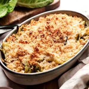 Baked cabbage casserole gratin topped with a golden, crispy breadcrumb crust in an oval dish, with cabbage leaves and a bowl of breadcrumbs nearby.