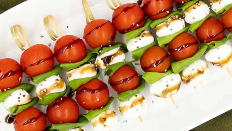 Skewers with cherry tomatoes, fresh basil leaves, and mozzarella balls drizzled with balsamic glaze, arranged on a white platter.