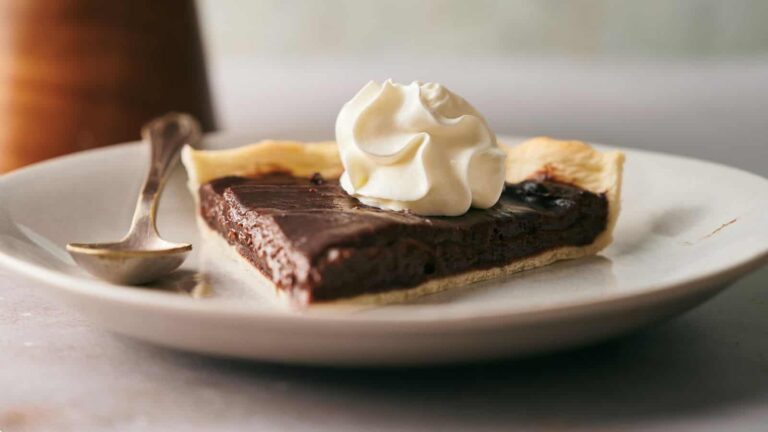 A slice of chocolate pie with a dollop of whipped cream on top sits on a white plate next to a spoon.