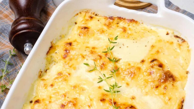 A baked potato gratin topped with melted, golden-brown cheese and garnished with fresh thyme in a white ceramic dish.