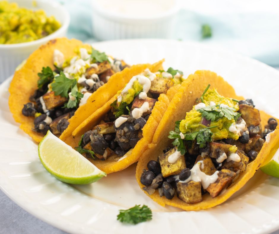 Three Sweet Potato and Black Bean Tacos filled with diced vegetables, guacamole, cilantro, and a drizzle of white sauce are arranged on a plate with lime wedges.