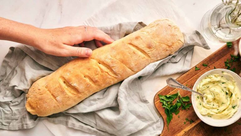 A hand reaches for a loaf of baguette resting on a cloth, next to a wooden board with herb butter and chopped parsley.
