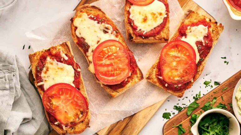 Four pieces of French bread pizza topped with tomato sauce, melted cheese, and fresh tomato slices on a parchment-lined wooden board. Fresh herbs are scattered nearby.