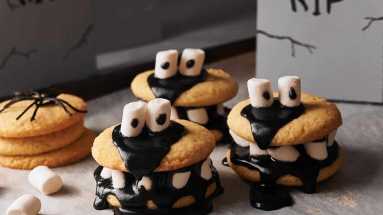Three sandwich cookies with black icing and marshmallow eyes and teeth are arranged on a table, resembling Halloween monsters. Additional cookies and marshmallows are visible in the background.