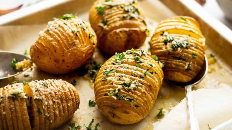 Five Hasselback potatoes topped with chopped herbs and grated cheese are arranged on a baking tray lined with parchment paper.