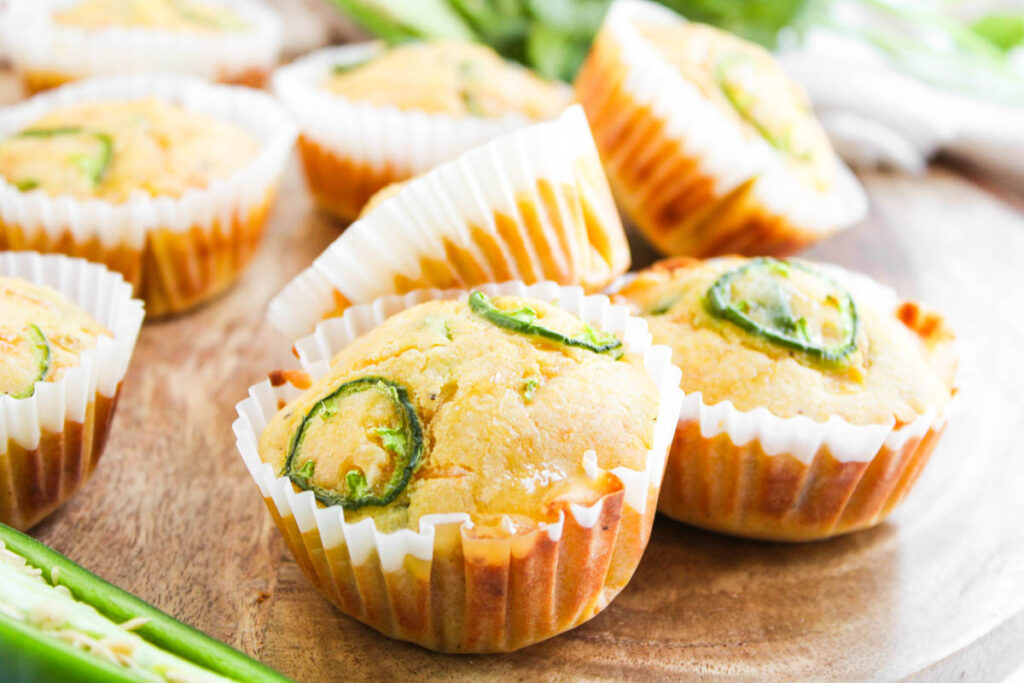 Three Jalapeño Honey Cornbread Muffins with jalapeño slices on top, in white paper liners, sit on a wooden surface with more muffins in the background.
