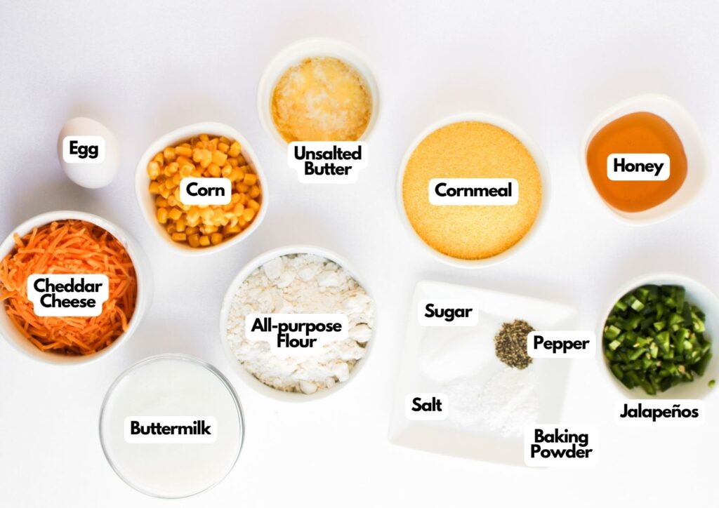 Top-down view of labeled ingredients for Jalapeño Honey Cornbread Muffins, including egg, corn, unsalted butter, honey, cornmeal, cheddar cheese, all-purpose flour, buttermilk, sugar, salt, pepper, baking powder, and jalapeños.