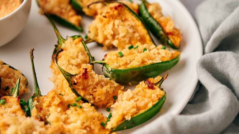 Halved jalapeños stuffed with a golden breadcrumb and cheese filling, garnished with chopped chives, arranged on a white plate next to a small bowl of dipping sauce.