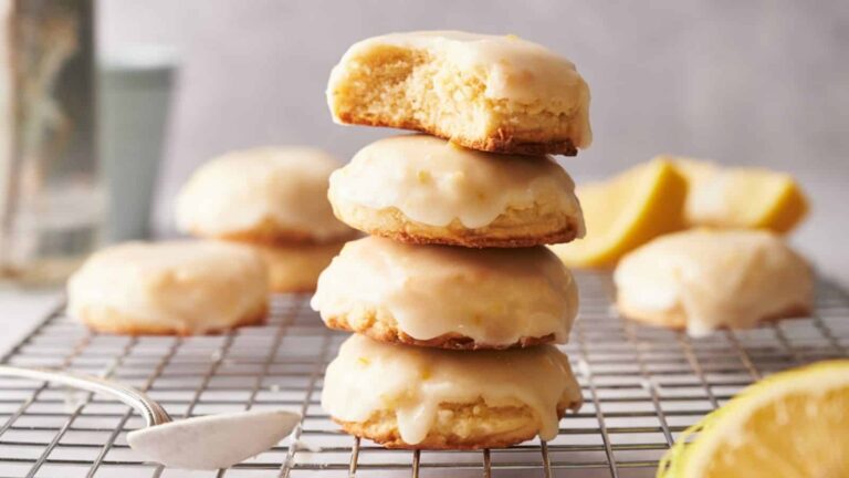 A stack of three glazed cookies sits on a cooling rack, with the top cookie partially bitten. More cookies and lemon slices are visible in the background.