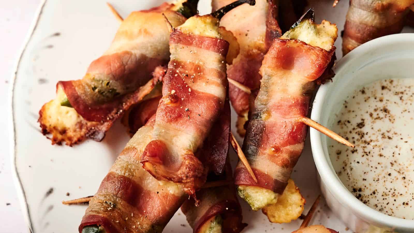 A plate of bacon-wrapped jalapeño poppers filled with cheese, secured with toothpicks, next to a small dish of dipping sauce.