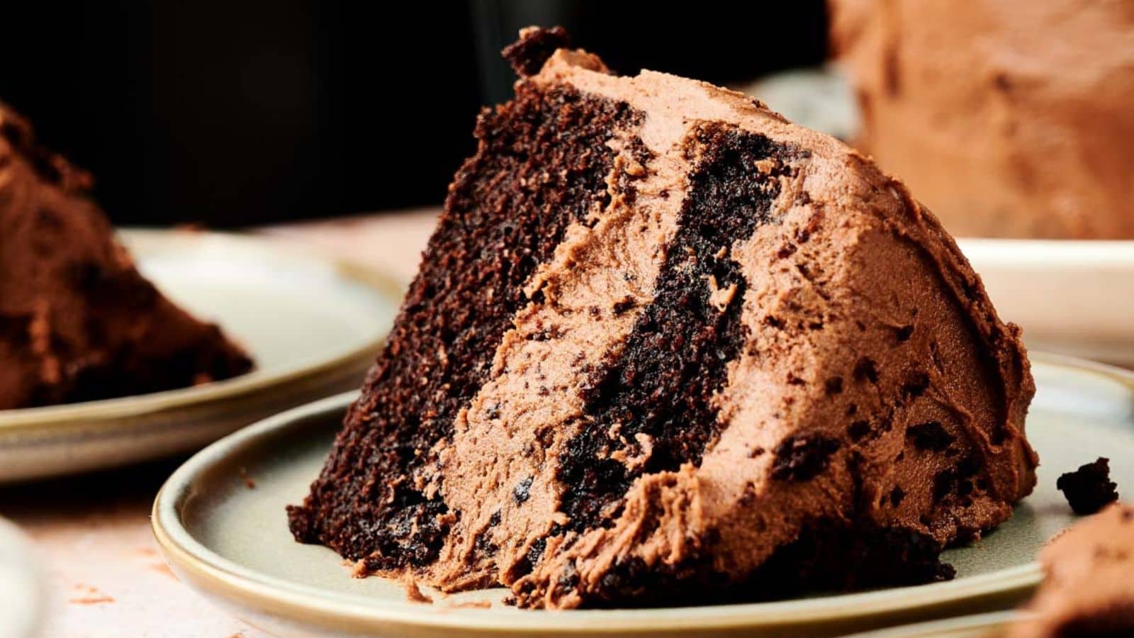A slice of chocolate layer cake with chocolate frosting is served on a beige plate.