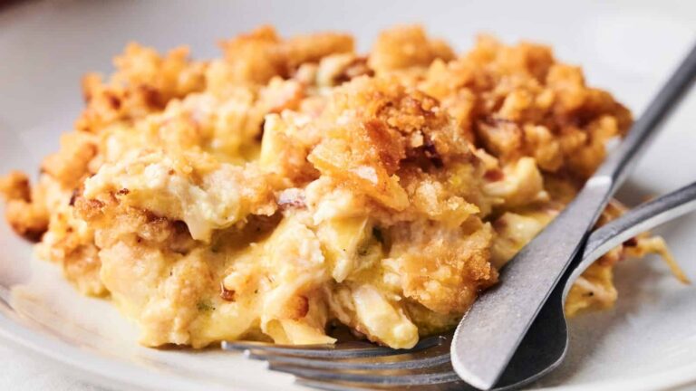 A serving of creamy casserole with a golden, crispy breadcrumb topping on a plate, with a fork and knife beside it.