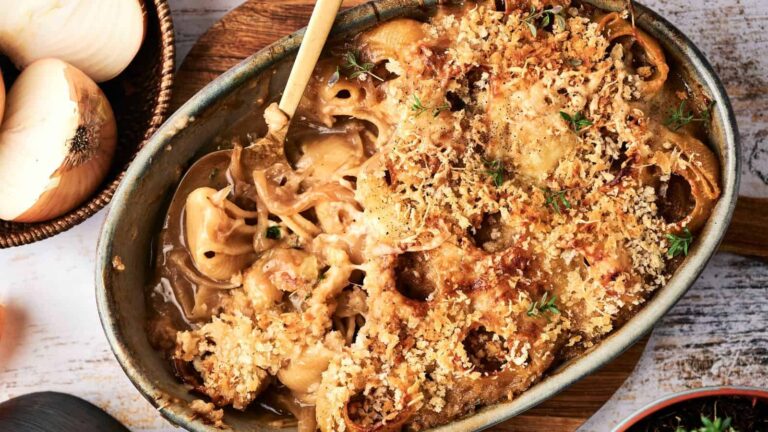 Oval baking dish with a golden-brown, breadcrumb-topped casserole, some served with a wooden spoon; onions and herbs visible nearby.