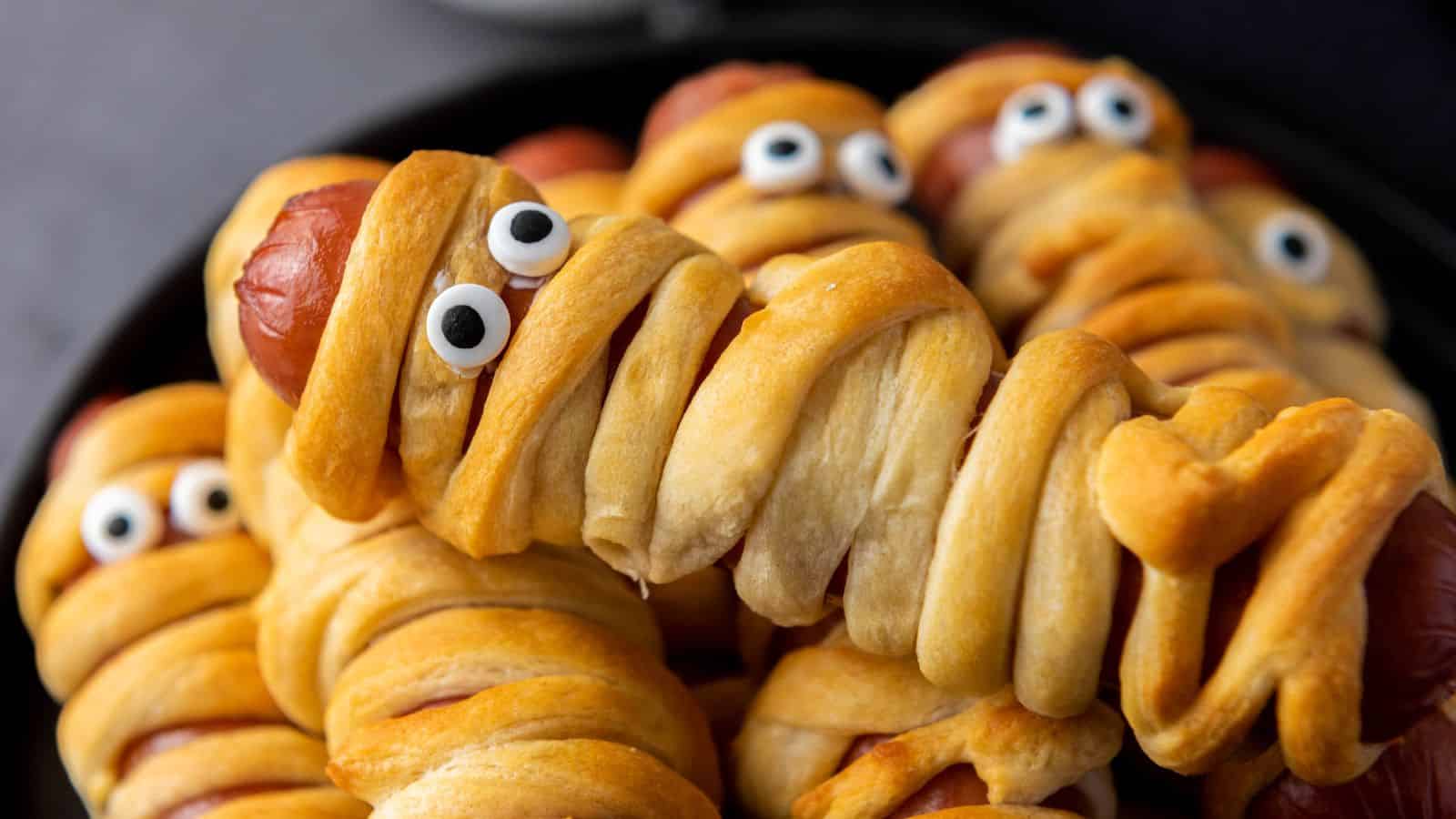 Hot dogs wrapped in dough to resemble mummies, complete with candy eyes, arranged on a black plate.