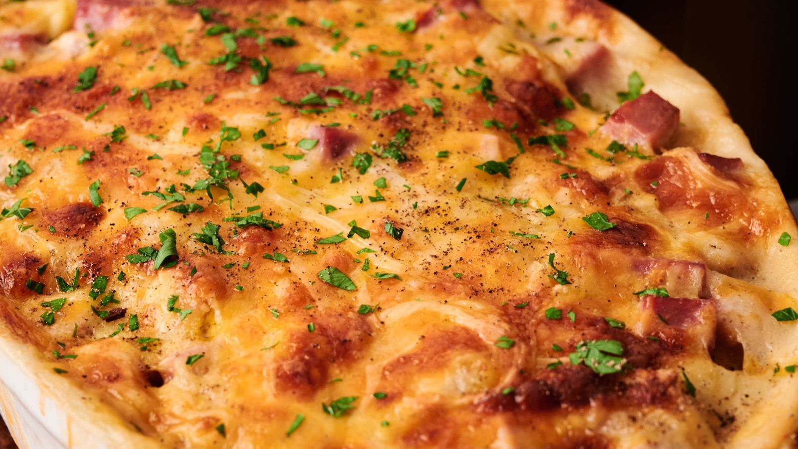 A close-up view of a baked casserole topped with melted cheese, diced ham, and chopped parsley.
