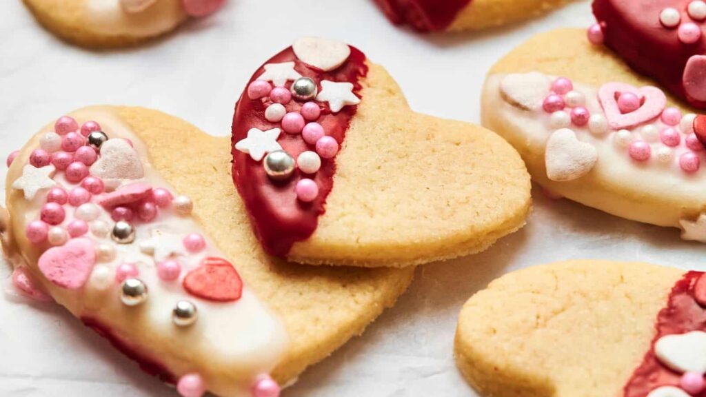 Heart-shaped sugar cookies decorated with red and white icing, sprinkles, and candy pearls, arranged on a white surface.
