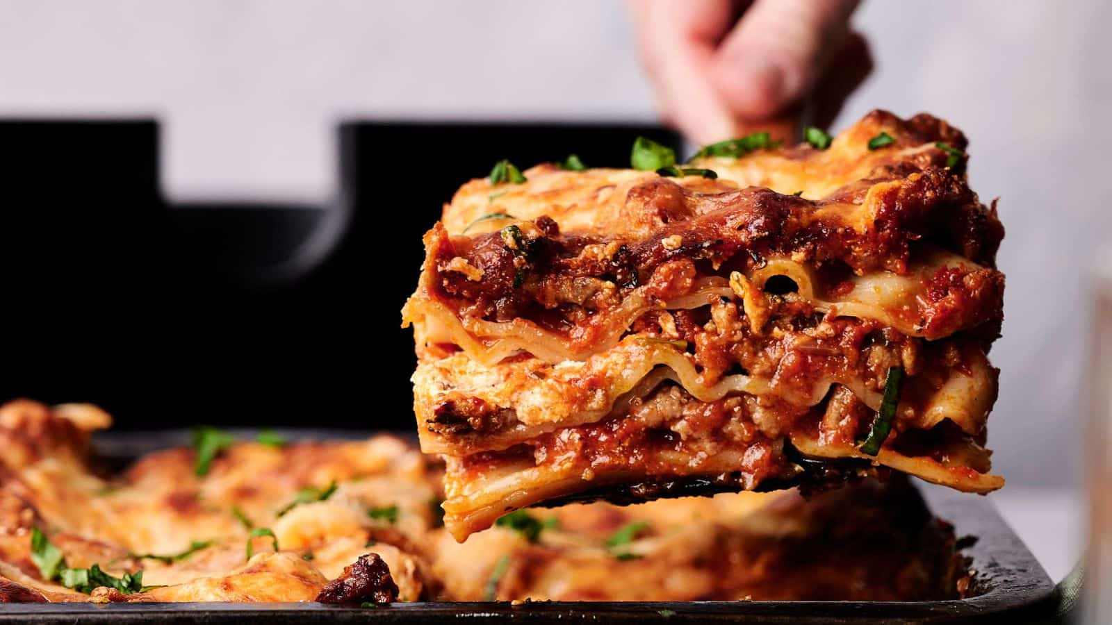 A close-up of a lasagna slice being lifted from a baking dish, showing layers of pasta, meat sauce, cheese, and garnished with herbs.