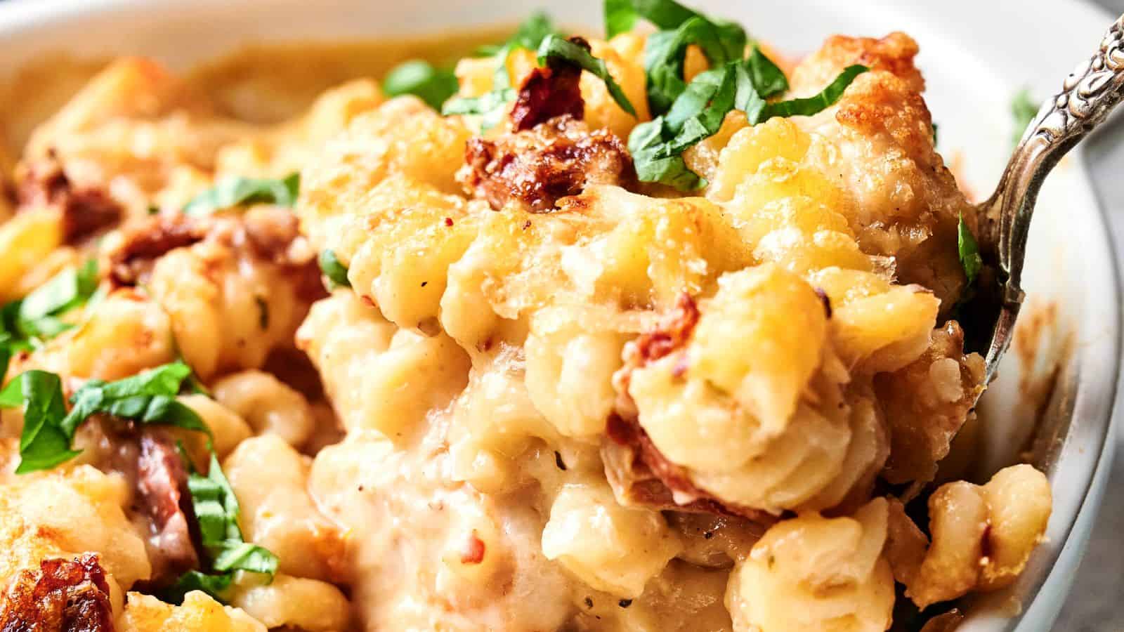 Close-up of creamy macaroni and cheese with crispy topping, garnished with chopped green herbs and pieces of sun-dried tomatoes.