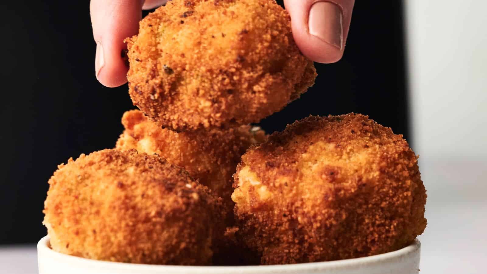 A hand picks a golden-brown, crispy fried ball from a bowl containing similar fried balls against a dark background.