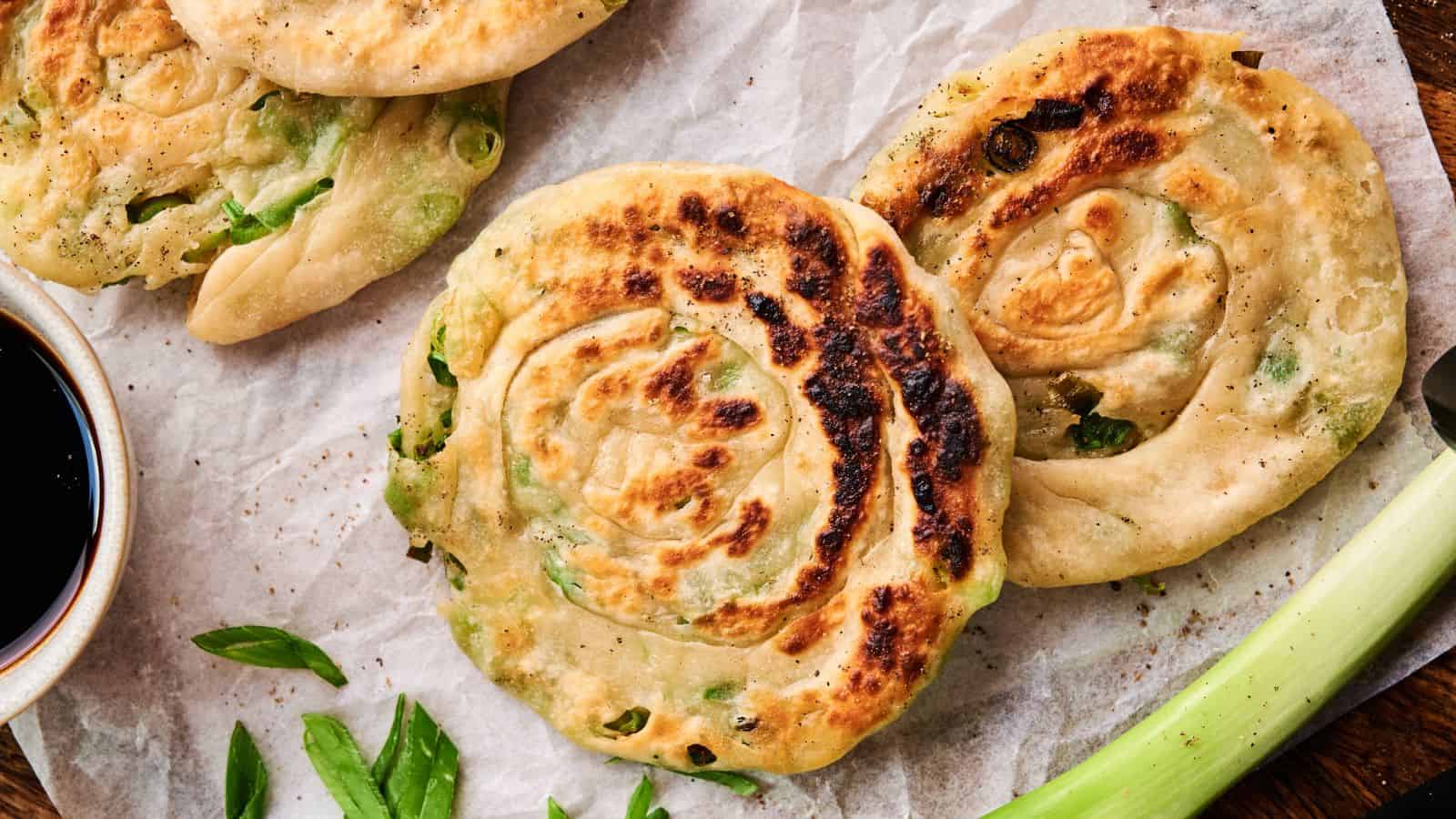 Three cooked scallion pancakes on parchment paper, accompanied by sliced green onions and a small dish of soy sauce.