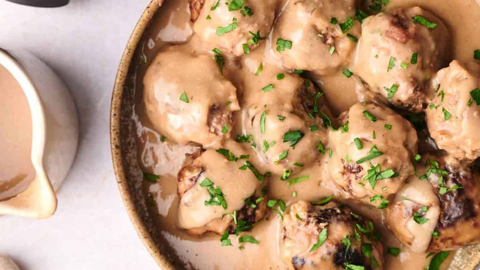 A bowl of meatballs covered in creamy gravy and garnished with chopped parsley, with a gravy boat to the side.