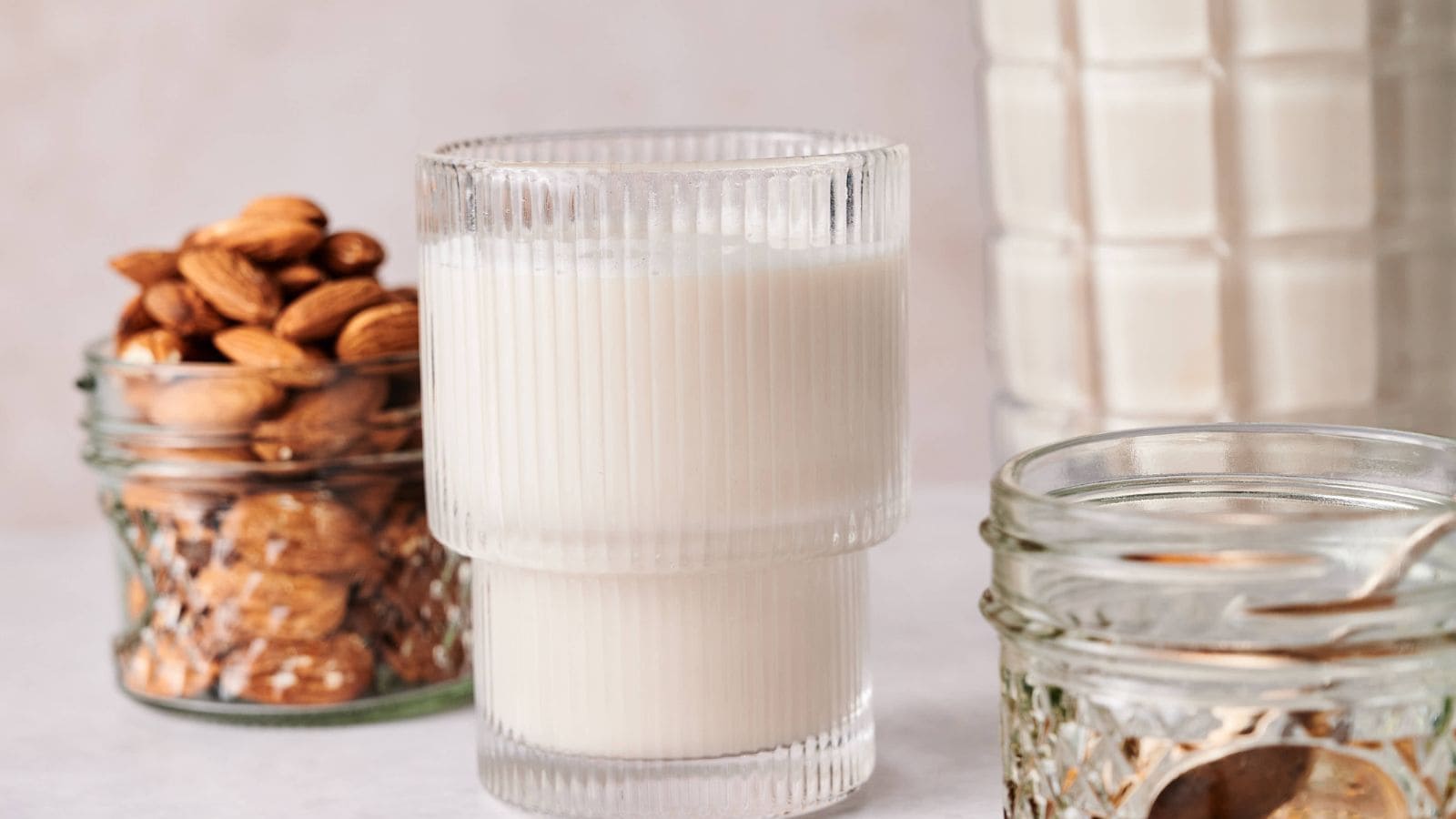 A glass of almond milk sits on a table next to jars containing whole almonds and other ingredients.