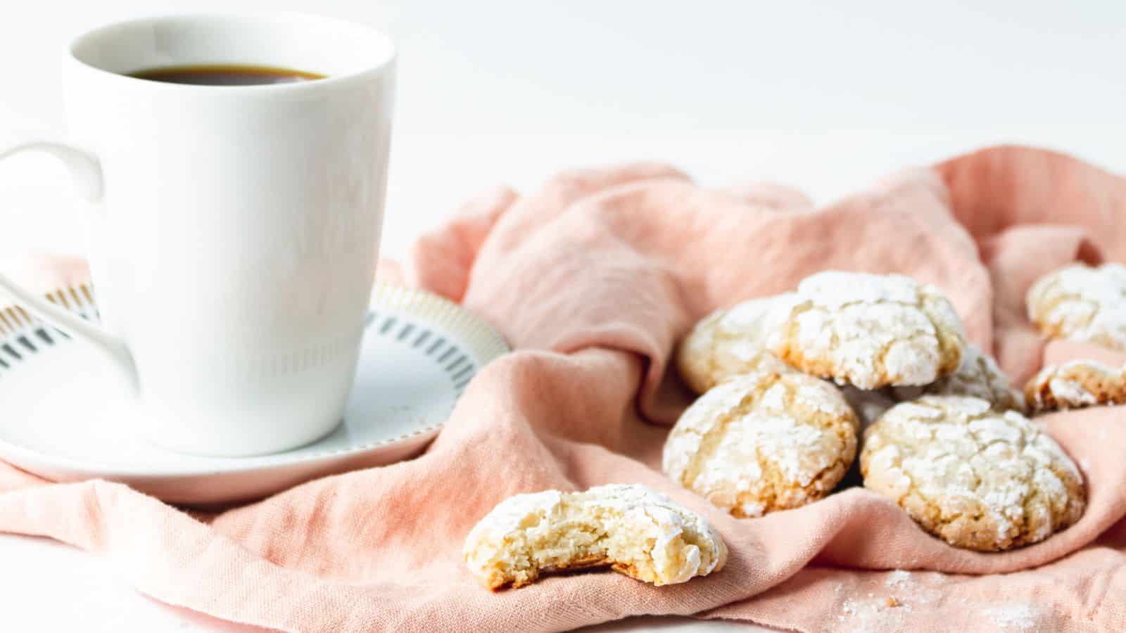 A white cup of coffee on a saucer sits next to several powdered sugar cookies on a pink cloth, with one cookie partially eaten.