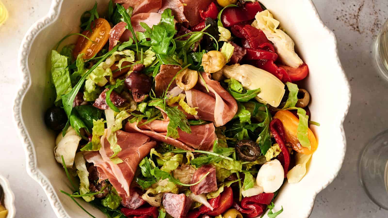 A bowl of salad with mixed greens, prosciutto, cherry tomatoes, olives, artichokes, and roasted red peppers.