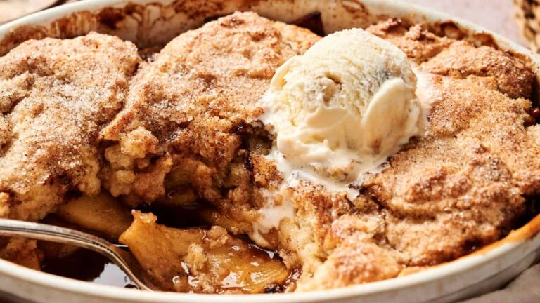A close-up of a baked apple cobbler topped with a scoop of vanilla ice cream, with a spoon resting in the dessert.