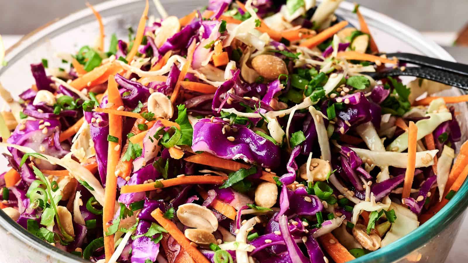 A bowl of colorful coleslaw with shredded purple and green cabbage, carrots, chopped peanuts, and greens, mixed with dressing.