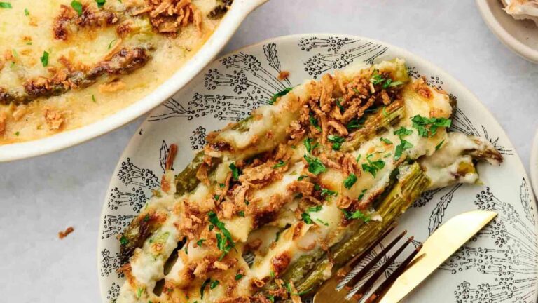 A plate with cheesy baked asparagus topped with crispy fried onions and chopped herbs, next to a gold fork. A casserole dish with more asparagus is partially visible.