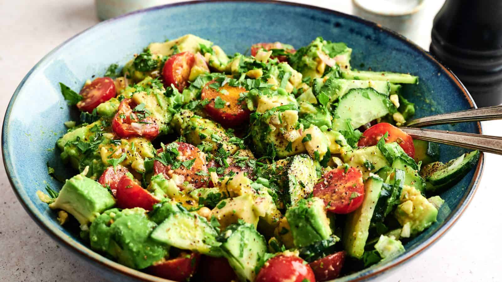 A blue bowl filled with a fresh salad containing avocado, cucumber, cherry tomatoes, herbs, and a light dressing, with a fork resting on the side.