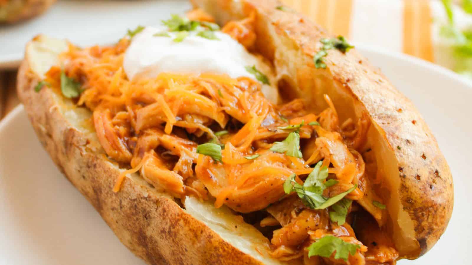 A baked potato topped with shredded barbecue chicken, sour cream, and chopped herbs on a white plate.
