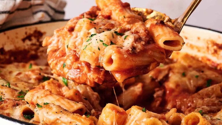 A serving spoon lifts baked ziti pasta covered in melted cheese and tomato sauce from a casserole dish.