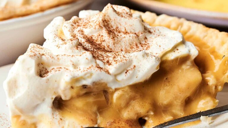 A slice of banana cream pie topped with whipped cream and sprinkled with cinnamon, with a flaky pie crust visible.
