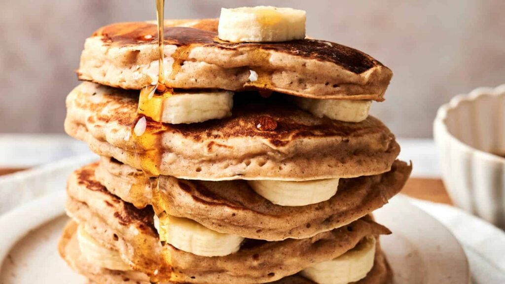 A stack of pancakes layered with banana slices is topped with more banana and drizzled with syrup.