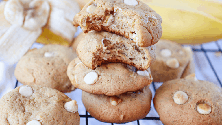 A stack of white chocolate chip cookies, with the top cookie partially bitten, sits on a cooling rack next to sliced bananas.