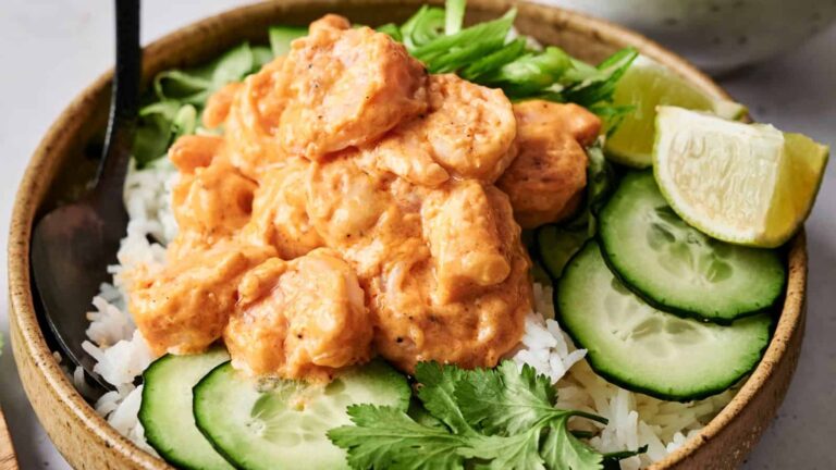 A bowl of white rice topped with creamy shrimp, sliced cucumbers, green onions, cilantro, and a lime wedge.