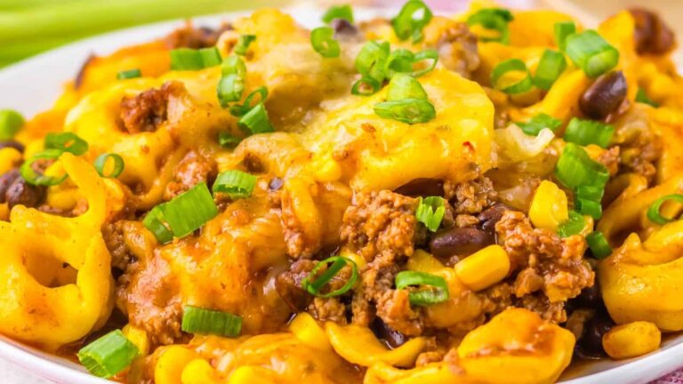 A close-up of a cheesy taco casserole with ground beef, black beans, corn, pasta, and chopped green onions on top.