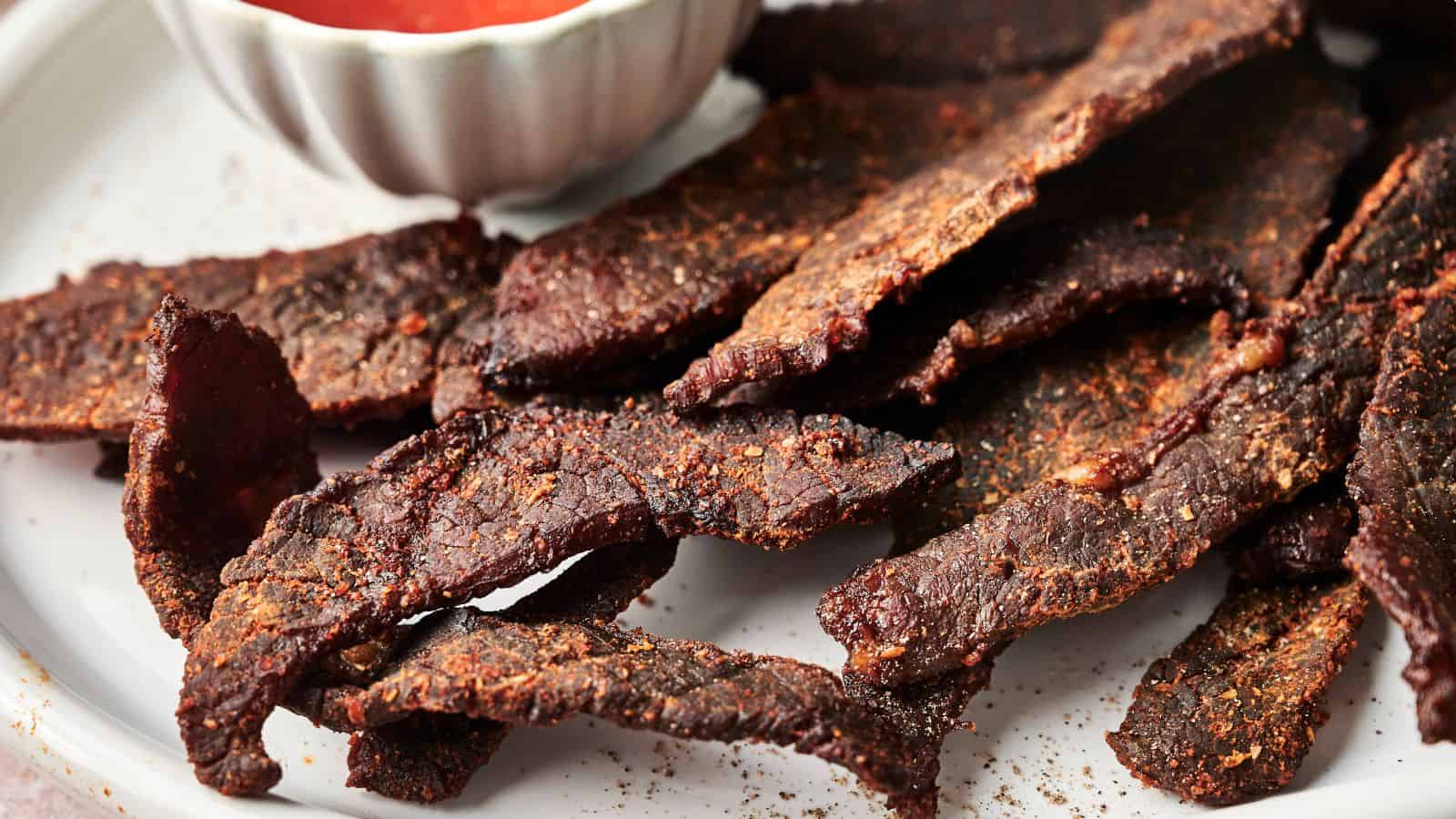 A plate of beef jerky slices next to a small bowl of dipping sauce.