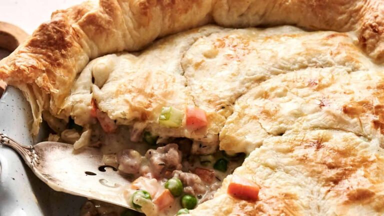 A close-up of a baked pot pie with golden, flaky crust, showing a filling of chicken, peas, carrots, and sauce, with a metal spatula lifting a slice.